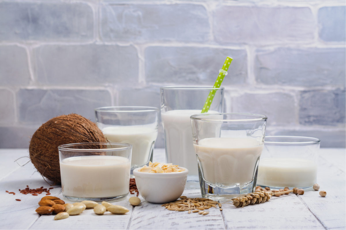 A selection of non-dairy milk in glasses, alongside a coconut and nuts. These are essential vegan baking ingredients.
