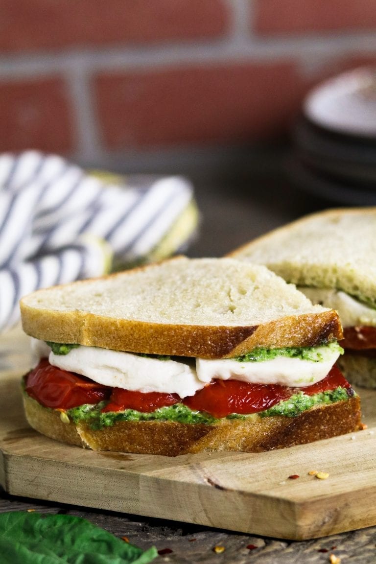 A vegan sandwich with thin artisanal bread is filled with a thick layer of vegan mozzarella cheese, roasted red peppers, and fresh pesto
