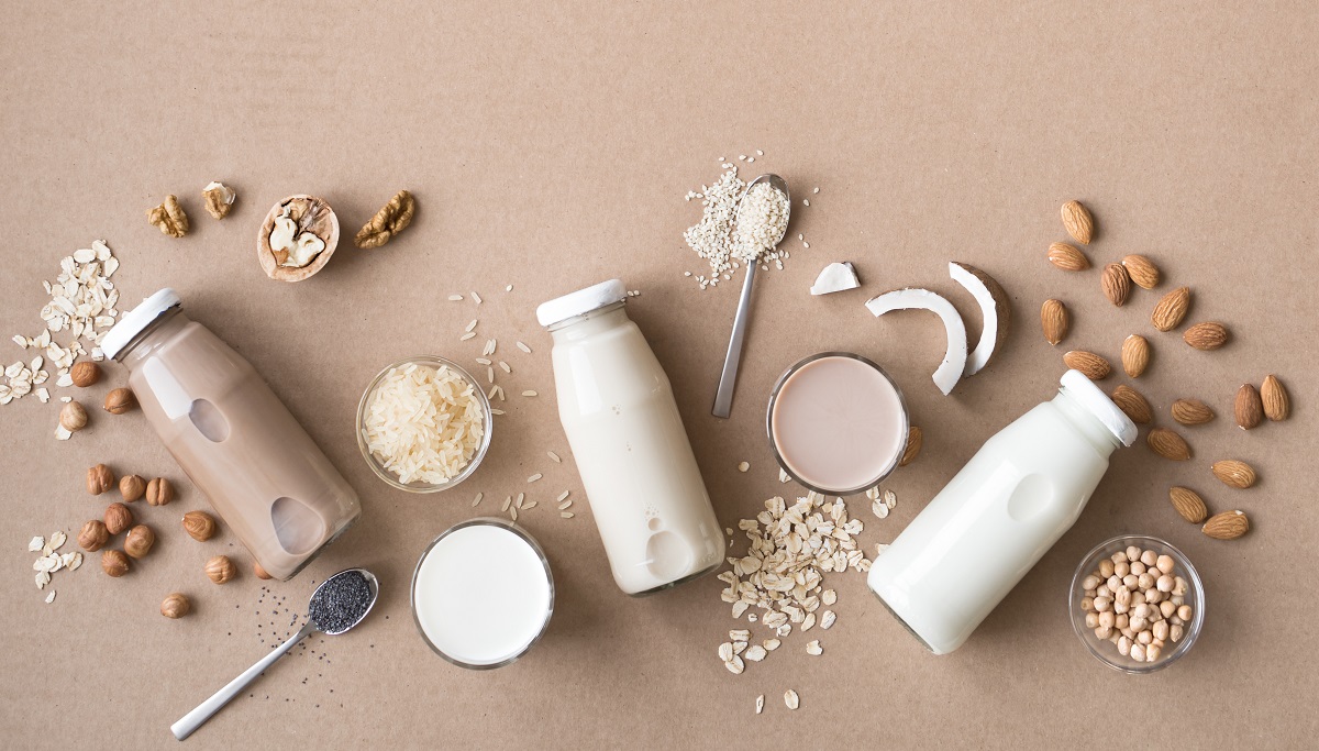 three glass jars of plant milks are surrounded by rice, oats, coconut slices, and nuts