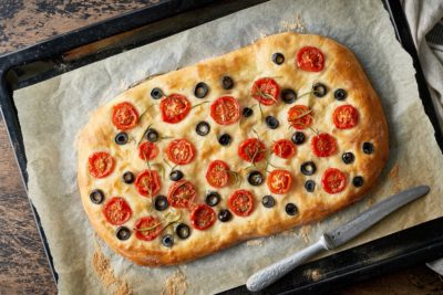 A delicious focaccia bread topped with olives, tomato slices, and rosemary sits on a piece of parchment paper