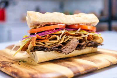 A fresh sub roll is stuffed with vegan meat slices, tomato, onion, and pepperocini