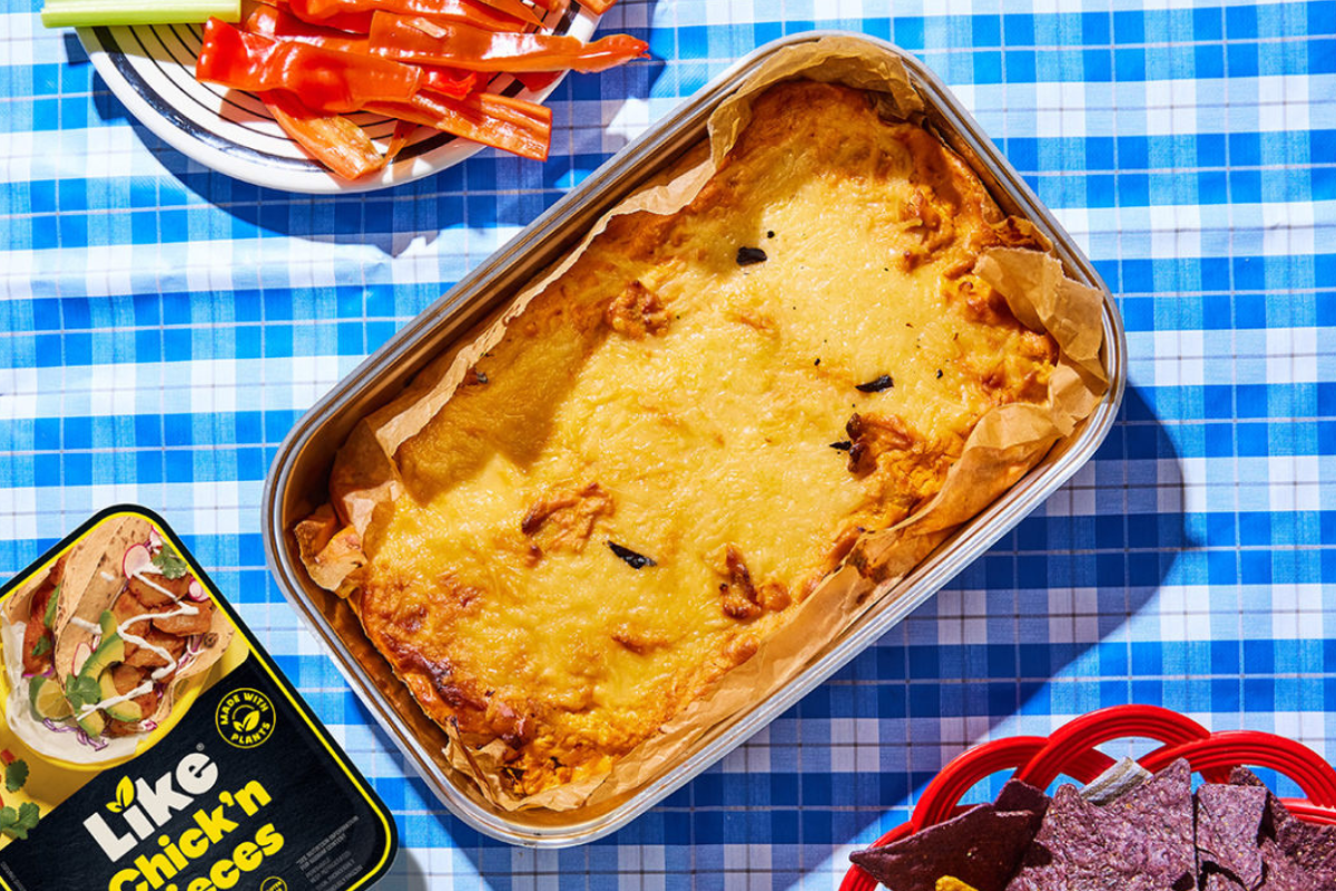 A pan of yummy vegan buffalo chicken dip is accompanied by a plate of veggies and tortilla chips on a bright blue gingham table cloth